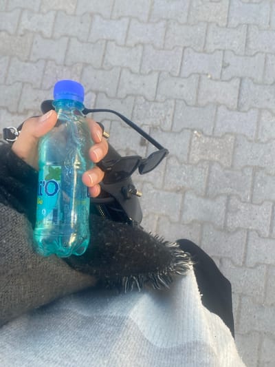 The image displays a close-up, top-down view of a person's lap or arm area, situated on a pedestrian walkway in Monastir, Tunisia. A light-skinned hand, featuring natural fingernails and a gold-colored ring on the ring finger, firmly grips a clear plastic bottle. The bottle has a blue cap, contains a light blue liquid, and bears a label with "O'LI" text and a green mint leaf graphic, indicative of a Tunisian mint-flavored beverage. Adjacent to the bottle, a pair of black, pointed-frame sunglasses rests on a dark-colored bag with visible gold hardware. The foreground includes dark, textured fabric with a frayed edge, layered over a lighter, possibly patterned fabric. The background consists of grey interlocking concrete pavers, characteristic of a sidewalk or public path.