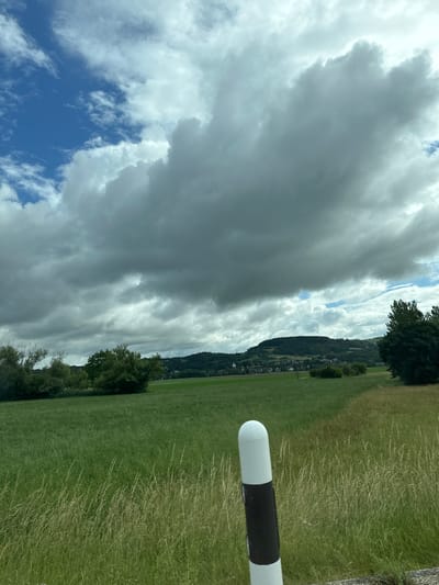 The image presents a wide, expansive landscape under a dynamic sky. In the foreground, prominently in the bottom right, a white cylindrical object with a rounded top is visible, featuring a distinct black band around its middle section, possibly a post or marker. The immediate ground consists of varied green grass, with some areas appearing longer and drier.

Beyond the foreground, a large, lush green field or meadow stretches towards a distant line of trees and hills. These hills are densely covered in dark green foliage, with hints of scattered buildings or houses nestled within, suggesting a rural or semi-rural settlement.

The sky occupies a significant portion of the upper frame and is marked by dramatic cloud formations. Large, heavy, dark grey clouds dominate much of the sky, giving an impression of overcast or possibly stormy weather. However, patches of bright blue sky are visible, particularly in the lower left and upper right corners, interspersed with lighter, wispy white clouds, indicating a changing weather pattern.

The scene captures a daytime moment with no people or specific activities taking place, focusing instead on the natural environment. The rolling, tree-covered hills and green fields under a compelling sky are consistent with the scenic landscapes found in areas like Zurzach, Switzerland. No visible text is present in the image.