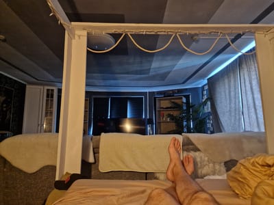 The image depicts a bedroom scene at night, as suggested by the low lighting and the illuminated television screen. The viewpoint is from a bed, looking towards a television and shelves in the background. In the foreground, a person's bare legs and feet are visible, resting on a peach-colored sheet. One foot is slightly raised, and the other is resting on a black sock with a red logo.
The room appears to be a living area or a multipurpose room, with a large television set as a focal point. To the right of the television, there are shelves displaying various items, including what appears to be a plant and decorative objects. The ceiling has a distinctive design with dark and light gray geometric patterns, and it's adorned with a rope and string lights, suggesting a decorative element or an attempt to create a specific ambiance. The room is furnished with a sofa or a large upholstered seating area draped with white faux fur rugs, and a beige throw blanket is draped over the back of a sofa. Curtains are visible in the background, drawn to the side.
The overall mood is relaxed and intimate, likely indicating a leisure activity like watching television or simply unwinding. The lighting is dim, with blue light emanating from the area around the television and curtains, and warm light from the string lights and possibly other hidden sources. There is no visible text other than the logo on the sock. The location context is Pogranichny, Russia.