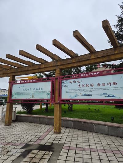 In the city of Hunchun, China, an outdoor scene features a wooden pergola structure with two large, rectangular display boards. The left board, with a light background and red border, contains Chinese text and an illustration depicting lotus flowers and a small dog. The right board, also with a light background and red border, has Chinese text and a landscape illustration of mountains, a body of water, and silhouetted figures. Both boards are mounted on yellow-painted pillars. In the background, a well-maintained green lawn is visible, dotted with several dark outdoor lights. Beyond the lawn, trees and a building with a red sign can be seen. To the left of the pergola, a small, three-wheeled vehicle and a trash receptacle are present. The ground in the foreground is paved with square tiles, some of which are wet, indicating recent rain or a puddle. The sky is overcast and gray.