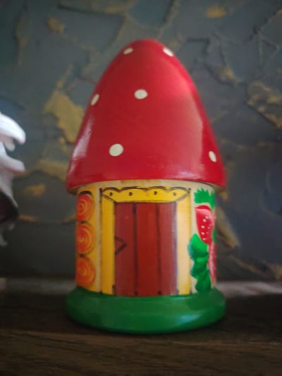 The image displays a close-up, slightly blurred view of a decorative mushroom-shaped house. The mushroom cap is bright red with white polka dots, tapering to a point. The base of the mushroom is painted yellow and features a red door with vertical lines and a yellow triangular accent. To the right of the door, there is a green painted strawberry with red seeds and a leaf. The sides of the yellow portion of the house have painted orange circular designs. The entire structure sits on a green, rounded base. The background is dark and textured, with hints of blue and yellow, and the object is placed on a dark, wooden surface. There is a blurry white object on the left side of the frame. The scene is indoors, likely on a shelf or table. There is no information or visual evidence to suggest this is located in the city of Сухарево, Russia.