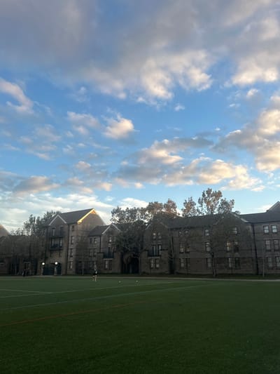 A low-angle view shows a college campus. A building with dark gray siding and many windows runs along the right side. In front of the building is a bright green athletic field. The sky is blue, with fluffy white clouds covering much of the frame. A few trees are visible between the building and the sky.