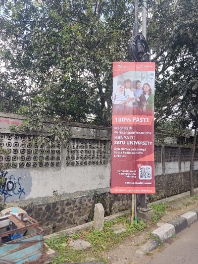 The image shows a street scene in Kecamatan Cimahi Selatan, Indonesia (latitude: -6.908, longitude: 107.567). A banner advertising "100% PASTI" internship and entrepreneurship opportunities at "SATU UNIVERSITY" is attached to a metal pole on the side of the road. The banner features four young adults smiling, with the text "Magang di Perusahaan/wirausaha HANYA DI SATU UNIVERSITY Ayo daftar sekarang! Kuota terbatas 100%" A QR code is also displayed on the banner. The pole is surrounded by green vegetation, and there are brick and concrete walls and a metal gutter behind it. A small vehicle is parked in the distance. The street is paved with gray asphalt and lined with white and black painted curbs. There are no pedestrians visible in the image.