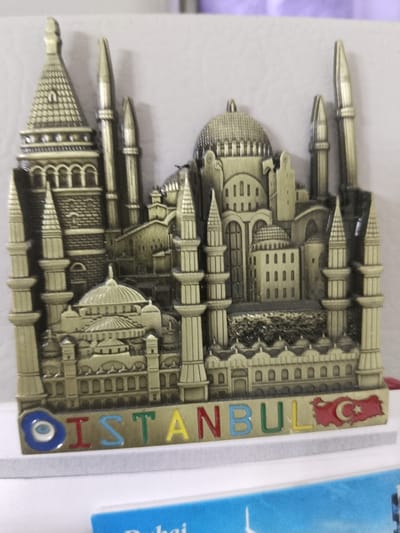 The image shows a souvenir magnet depicting the skyline of Istanbul, Turkey. The magnet is designed to resemble a metallic architectural model with intricate details of buildings and minarets. In the foreground, the word "ISTANBUL" is spelled out in colorful letters. To the right of the word, the flag of Turkey is depicted. The magnet appears to be placed on a white surface, possibly a shelf or table. In the background, a wall is visible, painted in a light, neutral color. There are no people or animals present in the image. The context of the city of Zhanaozen, Kazakhstan is not evident from the image itself.