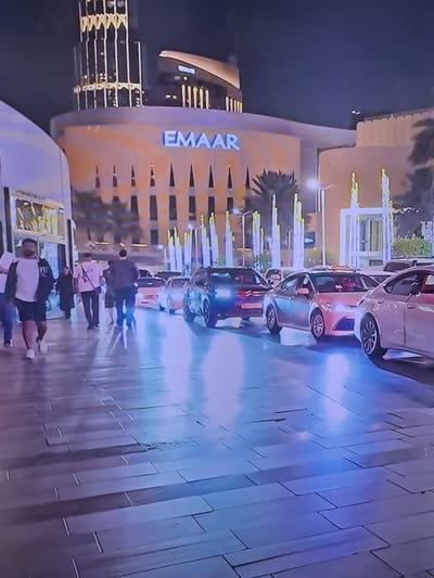 The media file shows an outdoor scene at night, with a modern building prominently featuring the blue text "EMAAR". The building has a curved facade with vertical light strips and windows. In front of the building, a row of cars is parked, and several people are walking or standing. The ground is illuminated by reflective blue and purple lights, suggesting it may be wet or made of a glossy material. The people appear to be casually dressed and walking in groups or individually. The overall atmosphere is one of an urban night setting, possibly a public space like a plaza or walkway outside a commercial or residential complex. The time of day is clearly night due to the dark sky and artificial lighting. There are no explicit weather cues, but the reflective ground could indicate recent rain or water features. The presence of the "EMAAR" text suggests a location associated with this company, which is a global real estate development company. The overall mood is relaxed and urban.
