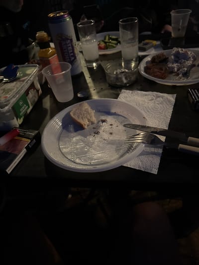 A table surface in Tankovo, Bulgaria, displays remnants of a meal and drinks. A white plastic plate in the foreground is partially empty, with scattered crumbs and a piece of bread. A fork and knife rest on a paper napkin to the right of the plate. To the left, a clear plastic container with green labeling, partially obscured, appears to hold a salad. Further back, a can of Stella Artois beer stands next to a tall glass containing a clear liquid, likely rakiya or ouzo. Another glass with a similar clear liquid is situated further back. A small glass ashtray filled with cigarette butts is visible to the left of the Stella Artois can. A bottle with an orange cap, possibly a condiment, is also present. A pack of cigarettes with red and white packaging lies to the left of the main plate. The lighting is dim, suggesting an evening or indoor setting. The presence of multiple drinks and plates implies a gathering of people, though only portions of individuals are visible in the background, out of focus. The table surface appears to be a dark, possibly metallic material.