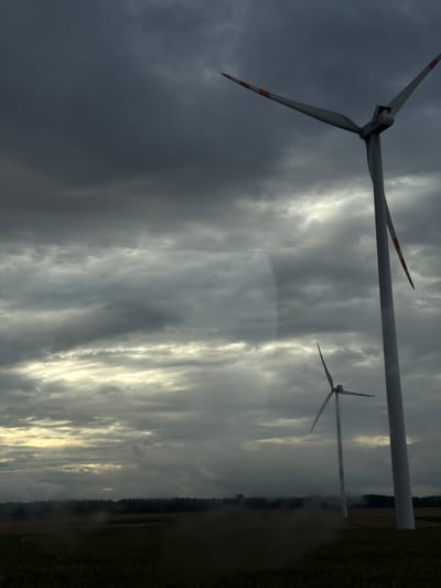 The image depicts a landscape with two wind turbines under a dramatic, cloudy sky. The foreground is a field, possibly farmland, with some texture suggesting grass or crops, partially obscured by a hazy or blurred effect, possibly due to rain or a dirty lens.
In the mid-ground, a forest line runs horizontally across the image, separating the field from the sky. The two wind turbines are the primary subjects. The larger, more prominent turbine is on the right, seen from a lower angle. Its white tower is tall and slender, and its three blades are angled upwards and outwards, with orange markings visible on the tips. The second, smaller turbine is further in the background, positioned to the left of the larger one, and appears to be the same design, with its blades also in a raised position.
The sky is filled with heavy, dark clouds, suggesting overcast or stormy weather. There are lighter patches in the sky, indicating some illumination, perhaps from a hidden sun breaking through the clouds.
The location is identified as Sieradz, Poland. The scene captures a moment of intense weather conditions over a rural or semi-rural area characterized by wind energy infrastructure. There are no visible people or animals. The focus is on the environmental and infrastructural elements of the landscape.