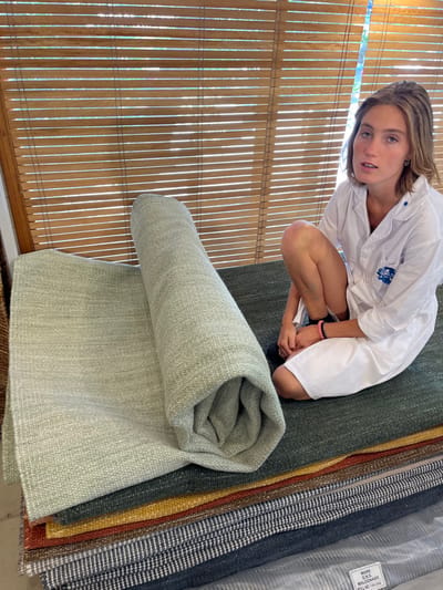 A person kneels beside a rolled light green rug and a stack of various colored rugs. The person wears a white shirt and has a bracelet on their right wrist. To the left, a bamboo shade filters sunlight. The stack of rugs features green, brown, yellow, striped, and grey patterns. A product tag is visible on the uppermost rug in the stack.