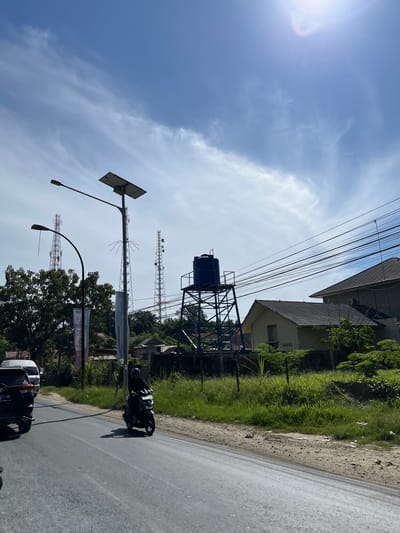 The image depicts an outdoor scene on a sunny day with a bright blue sky filled with wispy white clouds. The perspective is from a low angle, looking upwards and slightly to the side.

In the foreground, a paved road occupies the lower right portion of the frame. A black motorcycle, with a rider wearing a dark outfit and a helmet, is moving along the road. A portion of a black car is also visible on the road.

The middle ground features a grassy embankment on the left, with a dirt path running along its edge. Several structures are visible in the background. A tall, dark blue water tank sits atop a metal framework. To the right of the water tank, there are multiple telecommunications towers with various antennas. A street light with a solar panel is also prominent. Behind these structures, buildings with tiled roofs and lush green vegetation are present. Overhead, a complex network of electrical wires crisscrosses the sky.

The scene appears to be set in a semi-urban or rural area, possibly in Bukitrata, Indonesia, given the location context. The bright sunlight suggests it is daytime, likely mid-morning or afternoon, with clear weather. There is no discernible text visible in the image. The overall atmosphere is one of everyday activity, with traffic on the road and infrastructure contributing to the environment.