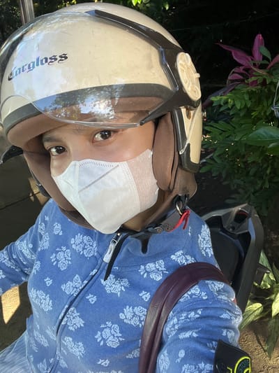 A person wearing a white face mask and a white "argloss" helmet, is positioned in front of green foliage and flowering plants. The person is wearing a blue zippered sweatshirt with white floral patterns and a dark shoulder bag. The scene appears to be located outdoors in Magelang, Indonesia.