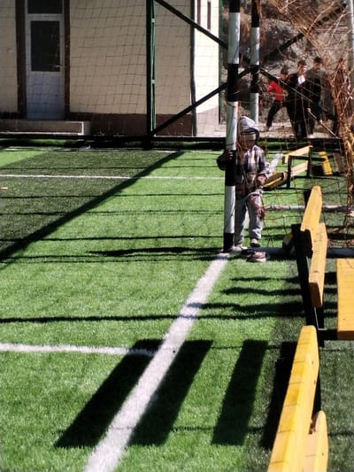The scene, located in the city of Limbur, Uzbekistan, depicts an artificial green turf sports field under direct sunlight, casting strong, elongated shadows. In the midground, a young individual, likely a child, stands centrally, clad in a grey plaid hooded jacket and grey pants, holding onto a black-and-white striped soccer goalpost. The hood is drawn, obscuring the face, with a light-colored band visible across the eye level. A tangled soccer net extends from the goalpost to the right. A pair of dark shoes and a light-colored object are positioned at the base of the goalpost, near the child's feet. To the right, a white and blue soccer ball rests on the turf adjacent to two partially visible yellow wooden benches. The background features a light-colored building with a dark door and window on the left. To the right, a metal fence with netting borders a barren, brownish hillside with sparse vegetation, where several indistinct individuals, appearing to be young males, are visible beyond the fence.