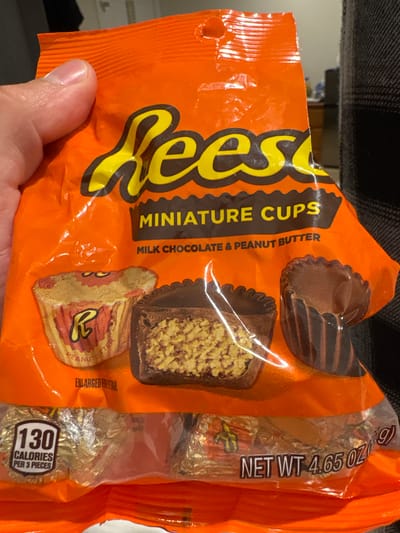 The image shows a hand holding a crinkled, orange-colored bag of Reese's Miniature Cups. The bag is made of a shiny plastic material, and its top left corner appears slightly torn or open, suggesting it has been handled or recently opened.
The dominant feature on the bag is the large, yellow, and brown "Reese's" logo. Below it, the text "MINIATURE CUPS" is visible, along with a description "MILK CHOCOLATE & PEANUT BUTTER". The packaging prominently displays illustrations of the candy: one whole Reese's cup, another cut in half to show the peanut butter filling, and a third unwrapped from its paper liner, revealing the rippled chocolate cup. Text below these illustrations reads "ENLARGED FOR DETAIL".
On the right side of the bag, a clear plastic strip reveals several individually foil-wrapped Reese's miniature cups inside. Vertical text on this strip indicates "NET WT 4.65 OZ (131g)". Further down, a white oval graphic with "130 CALORIES PER 3 PIECES" is clearly visible, providing nutritional information.
The hand holding the bag is in the lower-left portion of the frame, with the thumb partially obscuring some of the orange plastic. The background is dark and somewhat blurry, suggesting an indoor setting, possibly a piece of furniture like a couch or chair, given the textured fabric visible at the top. There are no other people visible, and no specific time of day or weather can be discerned from the image.