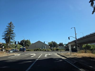 This media file depicts an outdoor scene at a street intersection under a clear blue sky, suggesting a sunny day. The view is from the perspective of someone on the road, looking forward. In the foreground, the asphalt road is marked with white directional arrows indicating lanes for going straight, turning left, and turning right.
The mid-ground features an intersection with traffic lights, some of which are illuminated green. Several portable restrooms are visible, placed on a paved area near the roadside. A white pickup truck is parked nearby. To the left, a tall evergreen tree stands prominently against the sky. Behind the portable restrooms and truck, a large, light-colored building with a distinctive triangular roof is visible. Further back, there is dense foliage and more trees.
In the background, a concrete overpass or highway bridge looms overhead, with cars visible on it. The setting appears to be urban or suburban, possibly in Sacramento, California, given the provided location context. The dry, golden grass along the side of the road hints at a warm climate. There is no indication of any specific activity or event taking place, beyond the normal flow of traffic at an intersection. The overall mood is neutral, reflecting a typical daytime scene. There is some text visible on a sign that says "ONE WAY" with an arrow pointing to the right. Another sign, partially obscured, appears to be a traffic regulation sign.