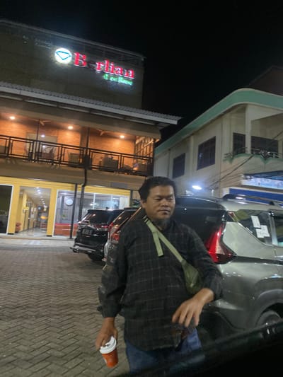 In this image, a man with a beard is standing next to a car at night. He is wearing a checkered shirt and a light-colored crossbody bag. In his right hand, he is holding a red cup with a white lid.
The setting appears to be an outdoor area with a brick-paved ground. To the left of the man, there is a building with illuminated signage that reads "Berkian Coffee". The building has a modern architectural style with balconies and windows. The time of day is clearly night, as indicated by the dark sky and the artificial lighting illuminating the scene. The overall mood seems casual and relaxed.