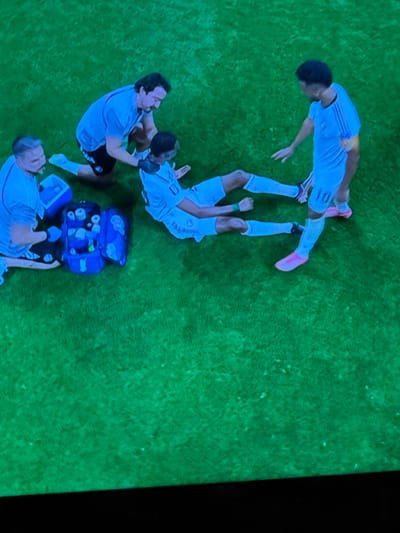 The image depicts a group of people on a grassy field, likely a soccer pitch, with a bright green hue. In the foreground, a person in a light blue jersey with the number "10" on the back is lying on the ground. Another person in a similar light blue jersey is kneeling beside them, appearing to offer assistance. A third person, also in a light blue uniform, is standing to the right, observing the situation. To the left, another individual in a light blue uniform is seated, and near them is an open blue medical bag filled with various items. The overall scene suggests a medical situation or injury assessment during a sporting event. The location is specified as the city of Bykovo, Russia.