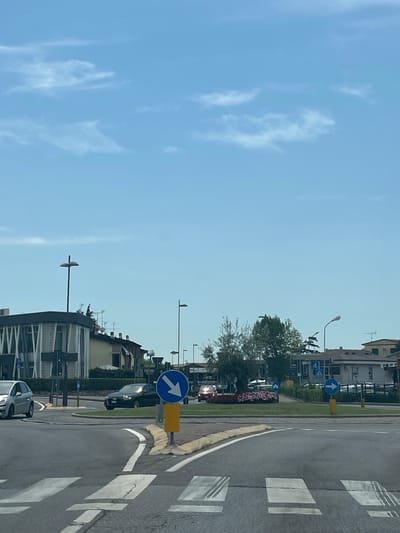 The image depicts a street scene in Castiglione delle Stiviere, Italy. The perspective is from a vehicle stopped at a crosswalk, looking towards a roundabout. The sky is clear with scattered white clouds.

On the left, a silver car is visible, partially obscuring a modern building with a white facade and horizontal louvers. A street lamp with a distinctive angled fixture stands in front of this building.

The roundabout features a circular patch of greenery with flowering plants and a small, gnarled tree. A dark sedan is positioned within the roundabout. A prominent blue circular traffic sign with a white downward-pointing arrow is mounted on a yellow pole to the right of the roundabout.

Further in the background, several residential buildings with tiled roofs are visible. Other traffic signs, including a blue circular sign with a white rightward-pointing arrow, are also present. Two other vehicles, one red and one black, are partially visible in the distance, navigating the roundabout. The road surface shows clear white markings for the crosswalk and lane dividers.