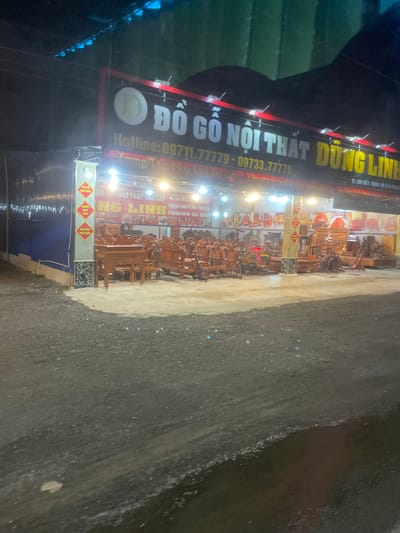 The image shows a nighttime view of a furniture store in Binh Thuan Ward, Vietnam. The store, named "ĐỒ GỖ NỘI THẤT DŨNG LINH," is illuminated by bright lights. A large sign above the store displays the name and contact information: "Hotline: 09711.77779 - 09733.77779." The store front is filled with various wooden furniture pieces, including chairs, tables, and intricate carved pieces. Some of the furniture is displayed on a paved outdoor area in front of the store, while more is visible inside through the open facade. Yellow banners with red text are attached to a pillar on the left side of the entrance, indicating "ĐỒ GỖ NỘI THẤT" and "GIẢM GIÁ 10%". The ground in front of the store is a rough, dark surface, possibly gravel or asphalt, with scattered small reflective elements, possibly due to moisture or lighting. The surrounding environment is dark, suggesting it is late at night.