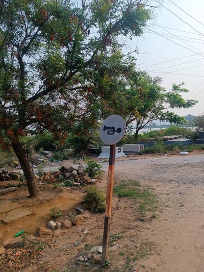The image shows a "Sound Horn" road sign in Kothacheruvu, India. The sign is round, gray, and has a black horn symbol on it. The signpost is orange and gray, standing on a dirt ground with rocks and sparse vegetation. Behind the sign, there is a tree on the left and a road leading to a cemetery or burial ground with concrete structures, likely tombs or memorials. There is also a body of water and hills in the background under a hazy sky. There is garbage strewn around the area, particularly near the tree on the left. Overhead power lines are visible.