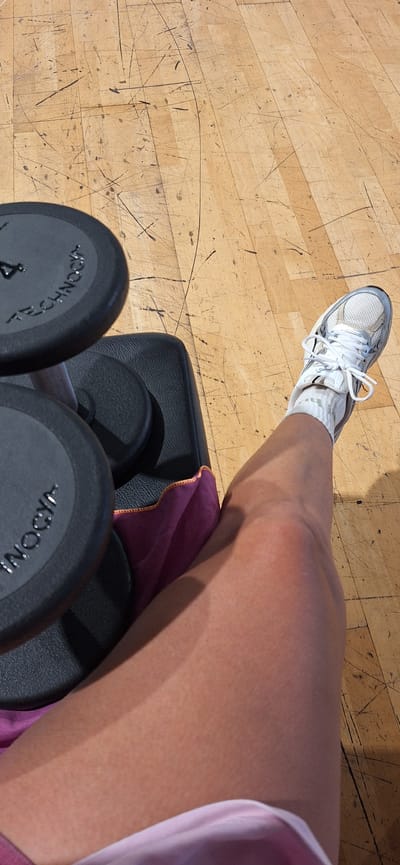 This image shows a close-up, high-angle view of a person's leg and foot in a gym setting. The person is wearing a white sneaker with white laces and a white sock. Their leg rests on a black fitness bench, partially covered by a small purple towel with an orange hem. In the foreground, a pair of black dumbbells are visible. On one dumbbell, the text "TECHNO" and the number "4" are clearly readable, indicating the brand and weight. The floor is made of light-colored wooden planks, heavily marked with numerous dark scuffs and scratches, characteristic of a well-used training area. Strong, distinct shadows suggest bright, direct lighting, likely indicating daytime. The scene, located in Santa Cruz De Tenerife, Spain, captures a moment of rest or preparation during a workout session.