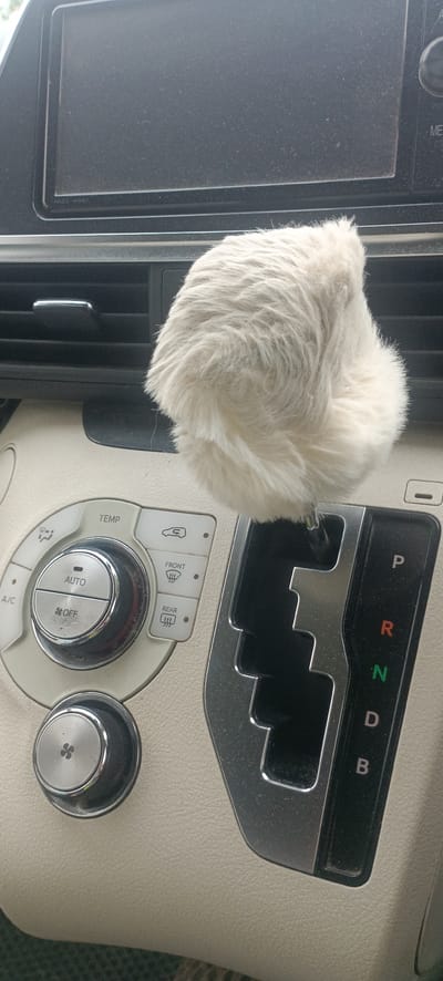 The image shows the interior of a car, focusing on the center console. A fluffy, cream-colored car accessory is positioned over the gear shifter. The gear shifter display shows "P", "R", "N", "D", and "B". To the left of the gear shifter are climate control buttons, including a large dial with "AUTO" and "OFF" settings, and smaller buttons labeled "TEMP", "A/C", and icons for defrosting the front and rear windshields. The dashboard is visible in the background, with a dark screen and air vents. The car's interior appears to be light-colored, possibly beige or off-white. The image is taken from a slightly high angle, looking down at the console.