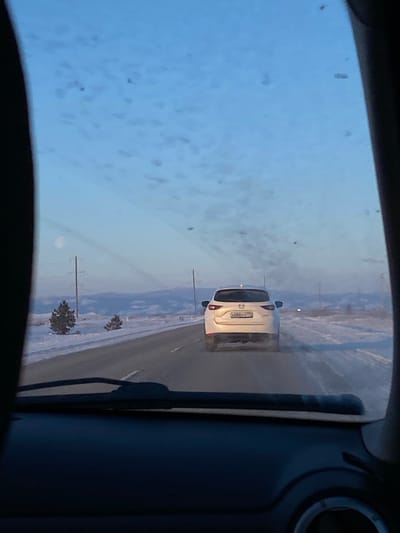 A view from inside a car looking out onto a snowy road. A white car with its license plate visible is directly ahead, traveling in the same direction. Snow covers the ground on either side of the road. The windshield is partially obscured by what appears to be water spots or ice. The car's interior is visible on the sides of the frame, with the dashboard and a partially visible air vent.