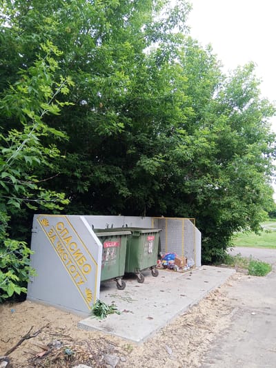 In Panino, Russia, a concrete slab area for waste bins is situated next to a dense line of green trees. A grey angled structure with yellow lettering saying "СПАСИБО ЗА ЧИСТОТУ" (Thank you for cleanliness) and a floral emblem is positioned on the left. Two large green trash bins with wheels are placed in front of the structure. A yellow metal mesh enclosure, containing various trash bags and items, is located to the right of the bins. The ground surrounding the concrete slab is covered with sand, debris, and scattered leaves. The image captures an outdoor scene, likely a designated recycling or refuse area within the town.