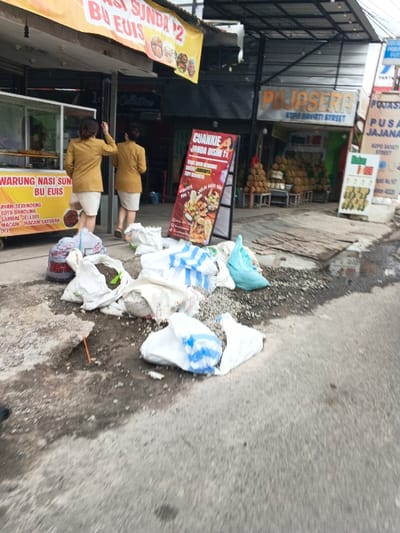 An outdoor scene showcases a local street vendor's food stall. The stall, "Warung Nasi Sun Bu Euis," offers various dishes, including Ayam Serundeng and Soto Bandung. In front of the stall, there are bags of trash. Two women in yellow jackets walk by, possibly customers. In the background, a durian vendor and another food stall "Pojasera" are visible. Street signage indicates "Kopo Sayati Street," further pinpointing the location. A colorful food advertisement sign stands on the sidewalk.