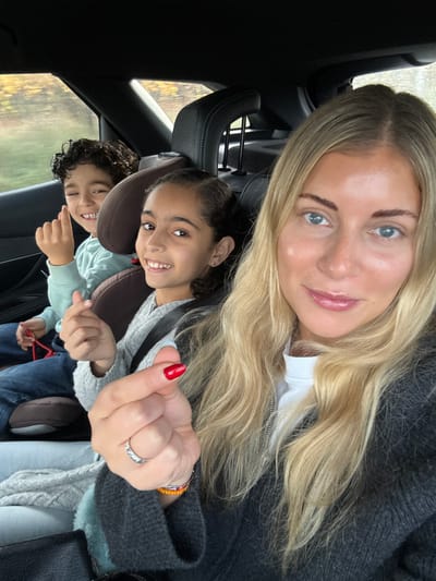 The image depicts three individuals inside a vehicle located in Pourrières, France, during daylight hours. In the foreground, an adult female with long, wavy blonde hair, light skin, and blue eyes is visible, facing slightly towards the camera. She wears a white high-neck top under a dark grey zippered cardigan. Her right hand is raised, forming a "finger heart" gesture, with the thumbnail painted red. A ring is present on her right ring finger, and beaded bracelets are visible on her right wrist. She displays a faint smile.
In the rear passenger area, two children are seated in booster seats. On the right, a girl with dark, pulled-back curly hair, light brown skin, and dark eyes, wearing a grey long-sleeved top, smiles directly at the camera. To her left, a boy with dark, curly hair, light brown skin, and dark eyes, wearing a light blue long-sleeved top and jeans, also smiles directly at the camera while performing a "finger heart" gesture with his right hand. A red object is partially visible near the boy's lap. The car's interior is dark, and blurred outdoor scenery, characterized by yellowish-brown foliage, is visible through the rear window.