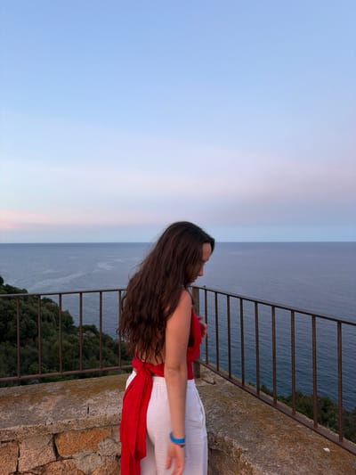 In the city of Santa Maria Navarrese, Italy, a young woman with long, dark hair stands on a stone balcony overlooking the sea. She is wearing a red top with a halter neckline and white pants. Her hair is partially obscuring her face as she looks down. The sea is a deep blue, stretching out to the horizon where it meets a pale blue sky with hints of pink and purple. A metal railing runs along the edge of the balcony, separating the woman from the view. Lush green trees are visible on the hillside below the balcony, adding a touch of nature to the scene. The overall impression is one of serene contemplation and natural beauty.