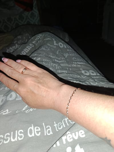 The image shows a person's left hand and forearm resting on a gray fabric with white text. The hand is adorned with a rose gold ring featuring a textured pattern and a delicate rose gold bracelet with small, diamond-like stones. The person has light-colored skin and manicured fingernails with a light pink hue and white tips. The fabric appears to be part of bedding or upholstery, with phrases in French visible, such as "bonjour," "matin," "l'imagination," and "rêves." The lighting is dim, suggesting an indoor setting, possibly a bedroom, and the overall scene is dimly lit, with the background blurred and dark. There is no identifiable information indicating a location in Vyatskoye, Russia.