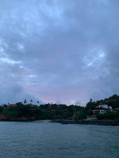 The image depicts a coastal scene at dusk in Batawala, Sri Lanka. The foreground is dominated by the ocean, its surface rippled and reflecting the muted colors of the sky. To the right, a rocky embankment separates the water from a lush, green hillside.

Nestled on the hillside are several structures, appearing to be resort buildings or villas. These buildings are predominantly white with some featuring visible balconies and lights indicating occupancy. The dense tropical foliage, including palm trees, covers the hillside, partially obscuring the structures and creating a sense of integration with the natural environment.

The sky above is filled with a heavy cloud cover, typical of tropical weather, and the light suggests the transition from day to night. Hints of pink and purple are visible near the horizon, indicating the presence of the setting sun behind the clouds. The overall mood is tranquil and serene, evoking a sense of a peaceful evening by the sea. There is no visible text in the image.
