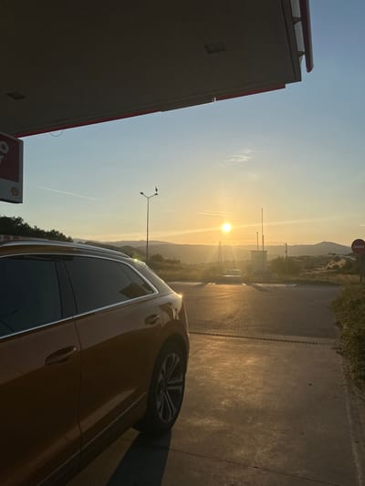 The image depicts a static scene at what appears to be a Shell gas station in Levunovo, Bulgaria, during either sunrise or sunset. A bronze or orange SUV, possibly an Audi Q8, is parked facing away from the viewer, with its rear passenger side visible in the foreground on the left. The vehicle's windows are tinted, and sunlight reflects off its body and roof rails.
Overhead, a large, dark-grey canopy of the gas station covers the immediate foreground, casting a shadow. A red stripe runs along the edge of the canopy. To the left, a portion of a red and white Shell sign is visible.
In the midground, the sun is low on the horizon, positioned centrally to the left, emitting a strong, golden light that casts long shadows across the concrete and asphalt pavement. The ground shows patches of illuminated concrete and darker asphalt. To the right, sparse vegetation and bushes line the edge of the pavement.
The background features a rural or semi-rural landscape with rolling hills or mountains. Various objects are scattered across the scene: a tall street light with a dark bird perched on its arm is visible behind the car's roof, slightly to the left of the sun. Further to the right of the sun, a set of utility boxes or small structures with antennae are present. A "No Entry" (red circle with white horizontal bar) road sign is prominently visible on the far right. A power transmission tower is faintly visible in the distance beneath the sun. The sky is clear, transitioning from a pale blue at the top to a golden hue near the horizon, with a faint contrail visible in the upper-left sky. No individuals are present in the image.
