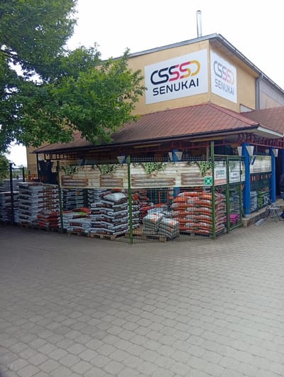 The image shows the exterior of a "Senukai" store in Kuršėnai, Lithuania. The store is a building supply and home improvement retailer. In front of the store, there is an outdoor display of bagged goods, likely soil or fertilizer, stacked on pallets and enclosed by a green metal fence. The bags are of various sizes and colors, with some clearly labeled. Signs are visible on the building and on wooden planks behind the fence, indicating product categories such as "TINKLAI" (nets), "LAUKO VAZONAI, TALPOS" (outdoor pots, containers), "SODO PRIEŽIŪROS PREKĖS" (garden care products), "DURPĖS" (peat), "SODO TECHNIKA" (garden technology), and "TRĄŠOS, ŽEMĖS" (fertilizers, soils). A sign for "5000 CENTRAS" is also visible. The building has a light yellow facade and a reddish-brown tiled roof. Trees with green foliage are present on the left side of the image, partially obscuring the building. The ground in the foreground is paved with interlocking grey bricks. A person is partially visible on the far right of the image, walking away from the store. The sky is overcast.