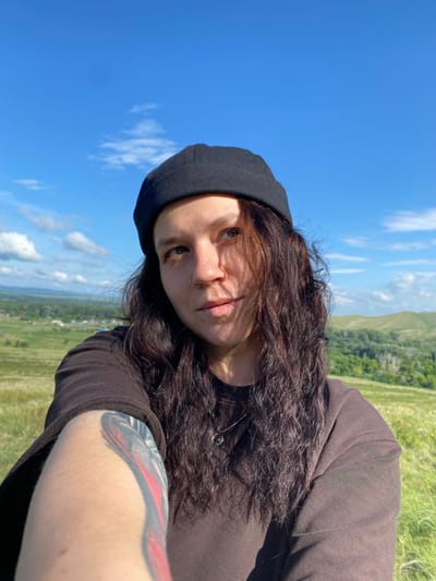 In a selfie taken outdoors, a person with long, dark, wavy hair and a black beanie is looking slightly away from the camera. They are wearing a dark brown long-sleeved shirt and have a nose ring. A colorful tattoo is visible on their left arm. The background features a lush green landscape with rolling hills under a bright blue sky with scattered white clouds. A small village or settlement is visible in the distance, nestled in the valley. The scene is set in Andreevka, Russia.