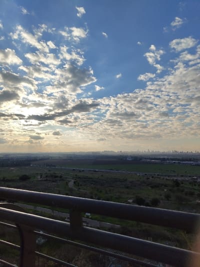 From a high vantage point, a view unfolds. A sky filled with scattered, white clouds stretches above. Below, a landscape reveals green fields intersected by a road and distant buildings. The sun is trying to break through the clouds. A portion of the image is obscured by the metal railing in the foreground. The scene evokes a sense of elevation.