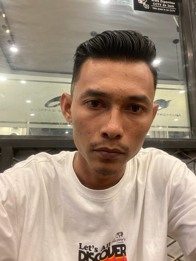 A man is shown in a close-up shot, presumably taken in Keudeublang, Indonesia. He has short, dark hair and is wearing a white t-shirt with text that includes "THE Discovery CHANNEL". The background reveals a shop window, possibly a coffee shop, with signage visible, including "COFFEE S" and "PREMIUM". A sticker on the window indicates "Area Dipantau CCTV 24 Jam," which translates to "Area Monitored CCTV 24 Hours."