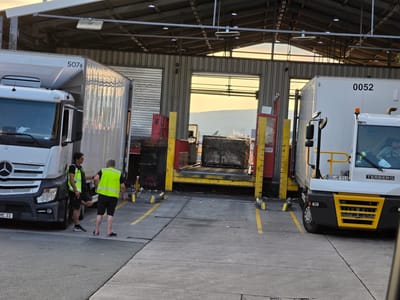 In Walldorf, Germany, two individuals are standing in a loading dock area. One person, wearing a high-visibility vest over a dark shirt and shorts, leans against a white semi-trailer truck. The other person, also in a high-visibility vest over dark clothing, stands facing the first person, with their left leg extended forward. To the right, a second white semi-trailer truck, identified by the license plate "GG HE 513", is parked. A forklift is visible at the loading dock, with a large object covered in plastic sheeting positioned on it. The scene takes place in front of a large building with a corrugated metal exterior and an open loading bay. The ground is paved with concrete and marked with yellow lines. The sky is visible in the background, appearing bright and possibly indicating sunset.