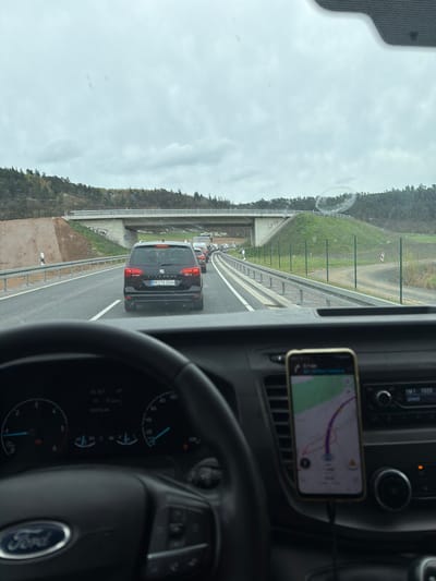 From the vantage point of the driver's seat of a vehicle, the scene depicts a traffic jam on a highway, likely in the vicinity of Göttingen, Germany, given the license plate “HR•TS 1014” visible on the rear of a black Seat Alhambra in the adjacent lane. A smartphone displaying a map is mounted on the dashboard. The environment includes overcast skies and wooded terrain along the roadside. The highway is elevated, with a bridge overhead. Several other vehicles are visible ahead in the traffic congestion. The Ford logo on the steering wheel is visible. No pedestrians are visible.