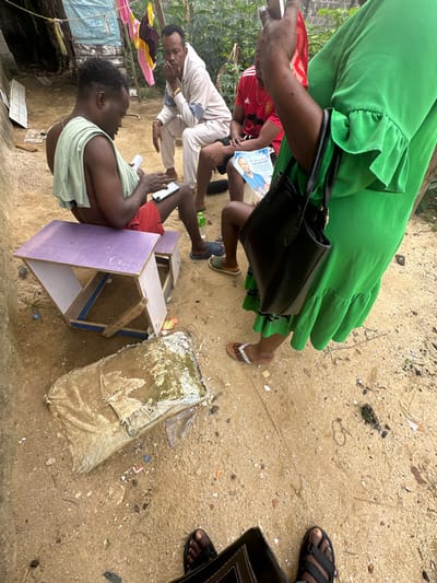 The scene captures an outdoor gathering of five individuals on a sandy dirt surface in Iriebe, Nigeria. In the mid-ground left, a dark-skinned male, shirtless with a light green towel draped over his shoulders, sits on a purple and white wooden bench. He wears red shorts and intently views a white smartphone held in both hands, with his left foot resting on a smaller matching stool. To his right, another dark-skinned male in a light beige hooded tracksuit sits, resting his chin on his right hand. Further right, partially obscured, a third dark-skinned male in a red Manchester United football jersey holds a light blue flyer featuring a portrait of a man and the word "Forever." In the immediate foreground right, a person wearing a green ruffled dress and brown flip-flops with white beaded straps stands, holding a black handbag with their left hand and a light-colored object, possibly a phone, in their right hand. The feet of the photographer, wearing black sandals, are visible in the lower foreground. The ground is unpaved, composed of light brown sand and small detritus, including a green plastic bottle and a rectangular, dirty, fabric-covered object. The background reveals a makeshift structure on the left, possibly constructed from metal sheets, with clothing hanging on a line, and dense green foliage beyond.