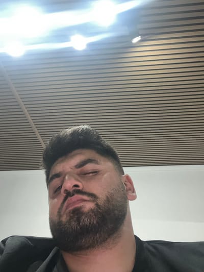 A close-up, low-angle selfie shows a man with dark, short hair and a full beard. He is looking down and to the left with his left eye partially closed, giving him a sleepy or weary expression. The man is wearing a black collared shirt. The background consists of a slatted ceiling made of light-colored wood, with bright overhead lights visible. The image appears to have been taken indoors, likely in an office or public space. No specific location in Rosenovo, Bulgaria can be identified from the image content.