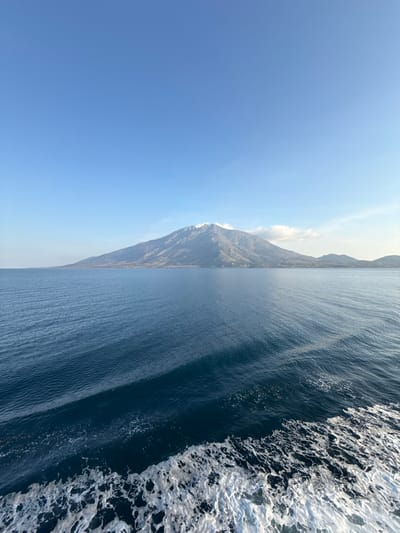 The scene presents an expansive view of the Aegean Sea and the island of Samothraki, Greece, observed from a moving vessel in the vicinity of Kamariotissa. The lower third of the frame consists of dark blue seawater, with a pronounced white foam wake concentrated in the bottom right, propagating from the viewpoint. Further out, the sea exhibits undulating ripples. Dominating the upper background is a large, rugged mountainous landmass, identified as Samothraki island, with its highest peaks displaying lighter grey rock or snow formations. The slopes are primarily dark green and brown, with a faint horizontal line of structures visible along the coastal base, indicating human habitation. The sky occupies the upper two-thirds, uniformly light blue and clear, with isolated small white clouds situated near the mountain's right peaks. No individuals or specific actions beyond the vessel's movement are discernible.