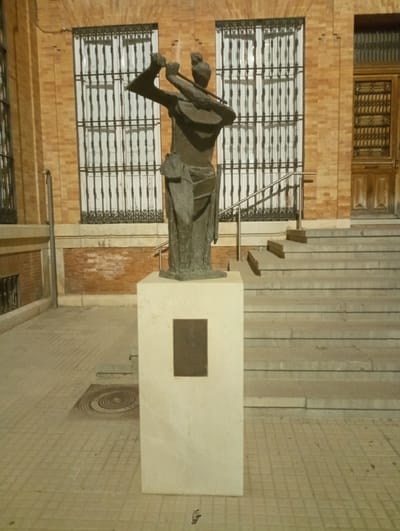 The image shows a bronze statue of a figure playing a stringed instrument, likely a guitar, standing on a light-colored stone pedestal. The statue is positioned outdoors in front of a building constructed of brick with decorative iron grilles on large windows. To the right of the statue, a wide set of stone stairs leads upwards. The ground in front of the statue is paved with square tiles. A circular decorative element is visible on the pavement to the left of the pedestal. The lighting suggests it is either dusk or nighttime. Based on similar images and art installations, this statue is likely located in Priego de Córdoba, Spain, possibly in a public square or near a cultural building.