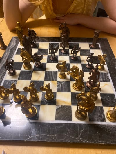 A marble chessboard displays an ongoing game. Gold pieces are arranged on the right side of the board, facing a darker set of pieces on the left. Both sides have pawns lined up, with a king and queen positioned at the back. Parts of two people are visible: one wearing a yellow dress sits on the left, and another person's arm is on the top left corner. The board is situated on a wooden surface.