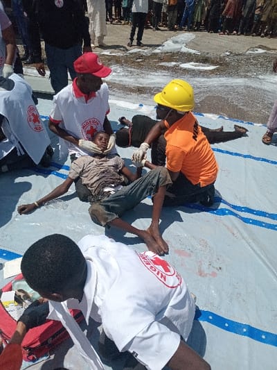 The scene takes place in Katabu, Nigeria, at the coordinates 10.71 latitude, 7.514 longitude. A young man is lying on his back on a white tarp with blue straps. His legs are bent at the knees, and his feet are together. A man in a yellow hardhat and an orange shirt is kneeling next to the young man, pressing on his chest, as if performing CPR. Another man in a red and white shirt, emblazoned with a red cross, is sitting behind the young man's head, his hands around the young man's neck. A fourth man, also wearing a white shirt with a red cross, is kneeling in front of the young man, reaching down to a red medical bag. There is a crowd of people in the background, some of whom are wearing white shirts with a red cross. The ground is covered with a white substance, possibly foam, or water mixed with dirt.