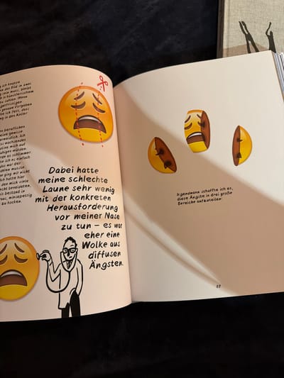 The image shows an open book with text and illustrations. On the left page, there is text in German and a large, sad emoji with tears. To the right of the emoji, there is a drawing of a person in a lab coat with a stethoscope, appearing to examine the emoji. On the right page, there are three illustrations of halves of sad faces, with text in German above them. The text describes emotions and anxieties. The overall scene suggests a humorous or metaphorical exploration of negative feelings. The image is a close-up of the book, with the surrounding environment out of focus. There is no information visible in the image that would indicate the location in the city of Mühlbach - Rio di Pusteria, Italy.