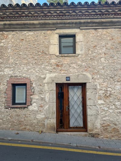 The image shows the exterior of a stone building with a tiled roof. There are two windows and a wooden door with glass panes. A house number, "66", is visible above the door. The building is located on a street with a sidewalk and a yellow line marking the edge of the road. Several fallen leaves are scattered on the sidewalk. The building appears to be in a state of disrepair, with some bricks missing around the smaller window. The sky is visible in the upper portion of the image.