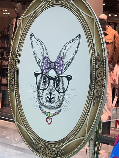 An oval-shaped display sign, approximately 1.5 meters tall, occupies the central foreground. It presents a monochromatic sketch-style illustration of a rabbit's head on a white background. The rabbit is depicted with prominent ears, whiskers, and a detailed snout. It wears a purple bow with white polka dots between its ears, black-rimmed eyeglasses, and a multicolored beaded necklace featuring a red heart pendant. The illustration is framed by an ornate gold-toned oval border with intricate scrollwork and a textured inner rim. The background, visible through or reflected on the sign's surface, shows an indoor retail setting. Two headless mannequins are discernible: one on the left wearing a collared shirt and dark tie, and another on the right in an orange-brown jacket and light-colored lower garment. Artificial plant elements, resembling birch branches and green foliage, are partially visible near the base of the sign. The floor appears to be light-colored and reflective. The scene is located in Vösendorf, Austria.