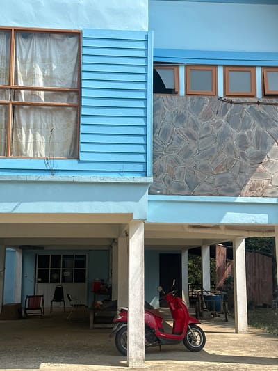 The scene is an exterior shot of a building in Ao Hin Khok, Thailand. The building has light blue siding and a stone wall facade. There are several windows, some with curtains visible inside. A red scooter is parked under a covered walkway supported by cream-colored pillars. Inside the walkway, there are chairs, a table, and what appears to be a dartboard hanging on the wall.