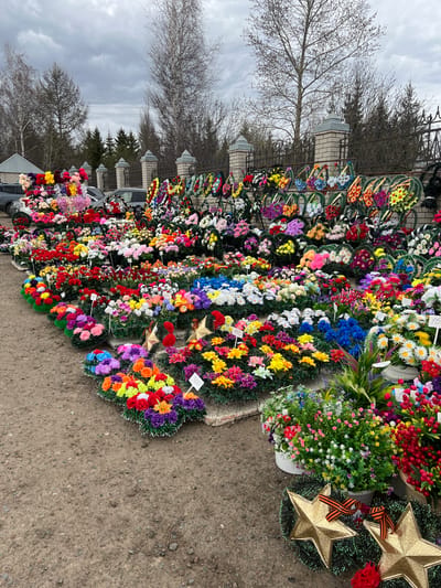 This image depicts a vibrant outdoor market stall or roadside vendor in Vysokaya Gora, Russia, specializing in artificial flowers and wreaths, commonly used for graves and memorials.

The primary subjects are a vast and colorful array of artificial floral arrangements. These include numerous wreaths of various shapes (circular, heart-shaped), individual bouquets, and larger potted arrangements. The flowers come in a wide spectrum of colors, including bright reds, yellows, blues, purples, pinks, whites, oranges, and greens, with common types resembling daisies, roses, and other small blooms.

Notable details include several wreaths shaped like gold stars, some of which are adorned with the distinctive orange and black striped St. George's ribbon, a symbol frequently associated with Victory Day commemoration in Russia. Small white price tags are visible on some wreaths, displaying numbers such as "150" and "300," likely indicating prices in Russian rubles.

The setting shows the floral display arranged directly on a paved or compacted dirt ground surface. In the background, a decorative metal fence with brick pillars runs behind the stall. Beyond the fence, several bare deciduous trees and some evergreen trees are visible, suggesting a late winter or early spring season. The sky is overcast and cloudy, indicating a daytime scene. Parts of a dark-colored car can be seen behind some of the flower arrangements, implying the location is near a road or parking area.

No people are visible in the image. The scene captures the organized display of goods for sale, reflecting cultural practices of remembrance and commemoration.