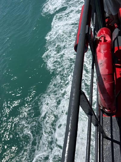 The image shows a view from the side of a moving boat, likely a ferry, looking out at the turquoise water of Thong Nian, Thailand. The water is choppy and creates a white wake trailing behind the boat. A dark metal railing runs along the side of the boat, with a red life preserver attached to it. The life preserver is cylindrical and appears worn. Sunlight glints off the water, creating many bright reflections. In the upper right corner of the image, a person's legs, clad in white, are visible, suggesting someone is standing near the railing. The scene is captured from a high angle, looking down and slightly forward.